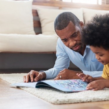 Dad is reading a book to his son while laying on a white rug on the floor.