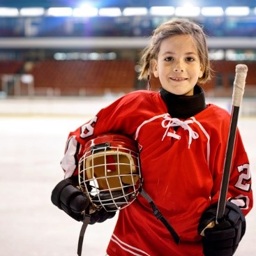 Child dressed in red hockey uniform holding a hockey mask and hockey stick while standing on an ice rink.